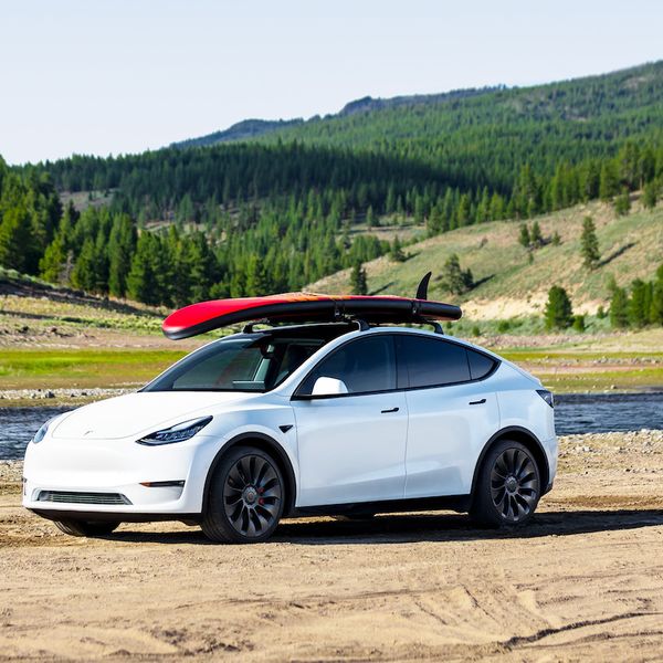 Tesla Model Y with a paddle board on the roof rack. White car, on a beach in mountain area
