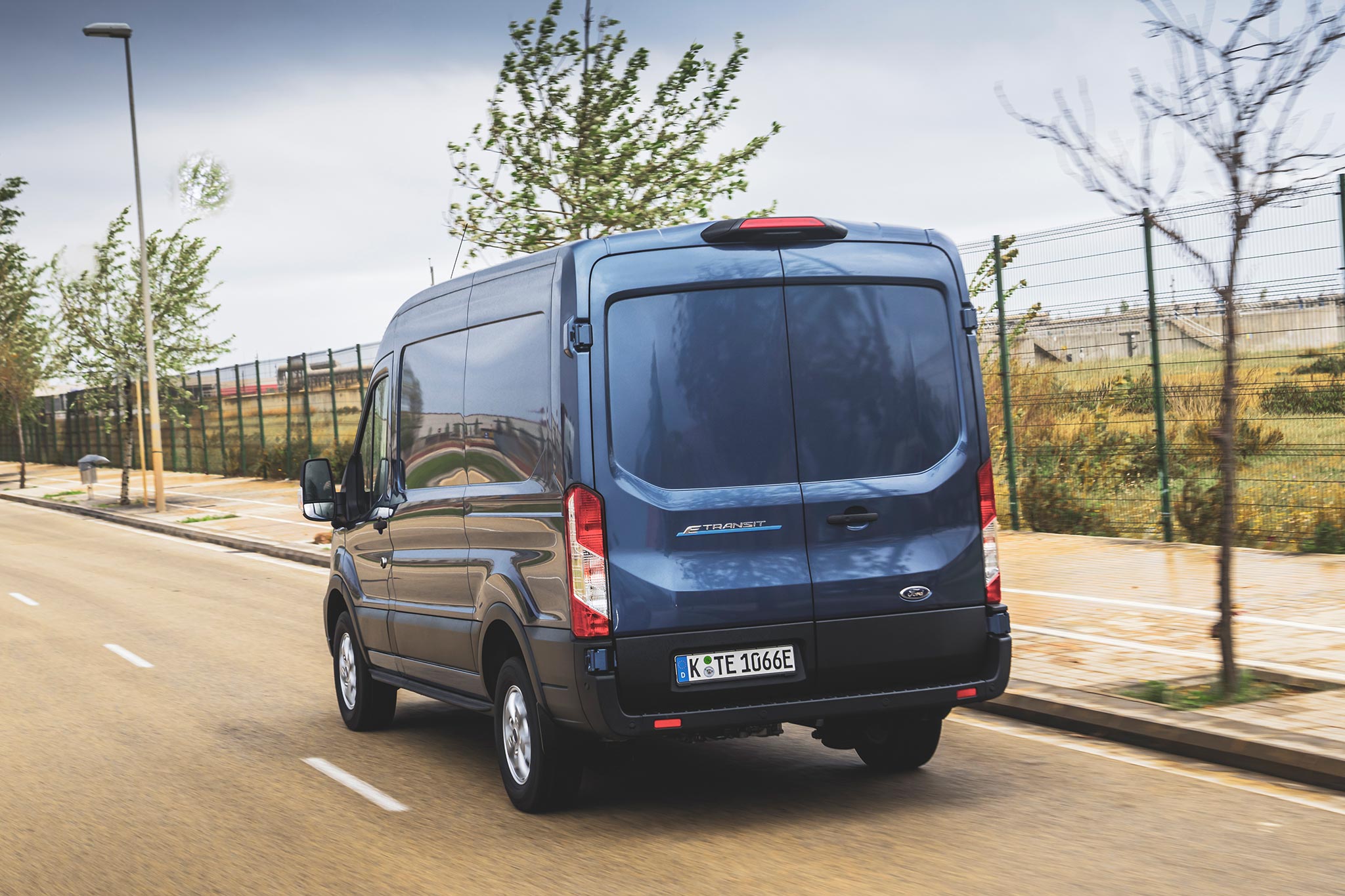 Ford E-Transit in Chrome Blue, German registered, rear tracking action shot in front of fence